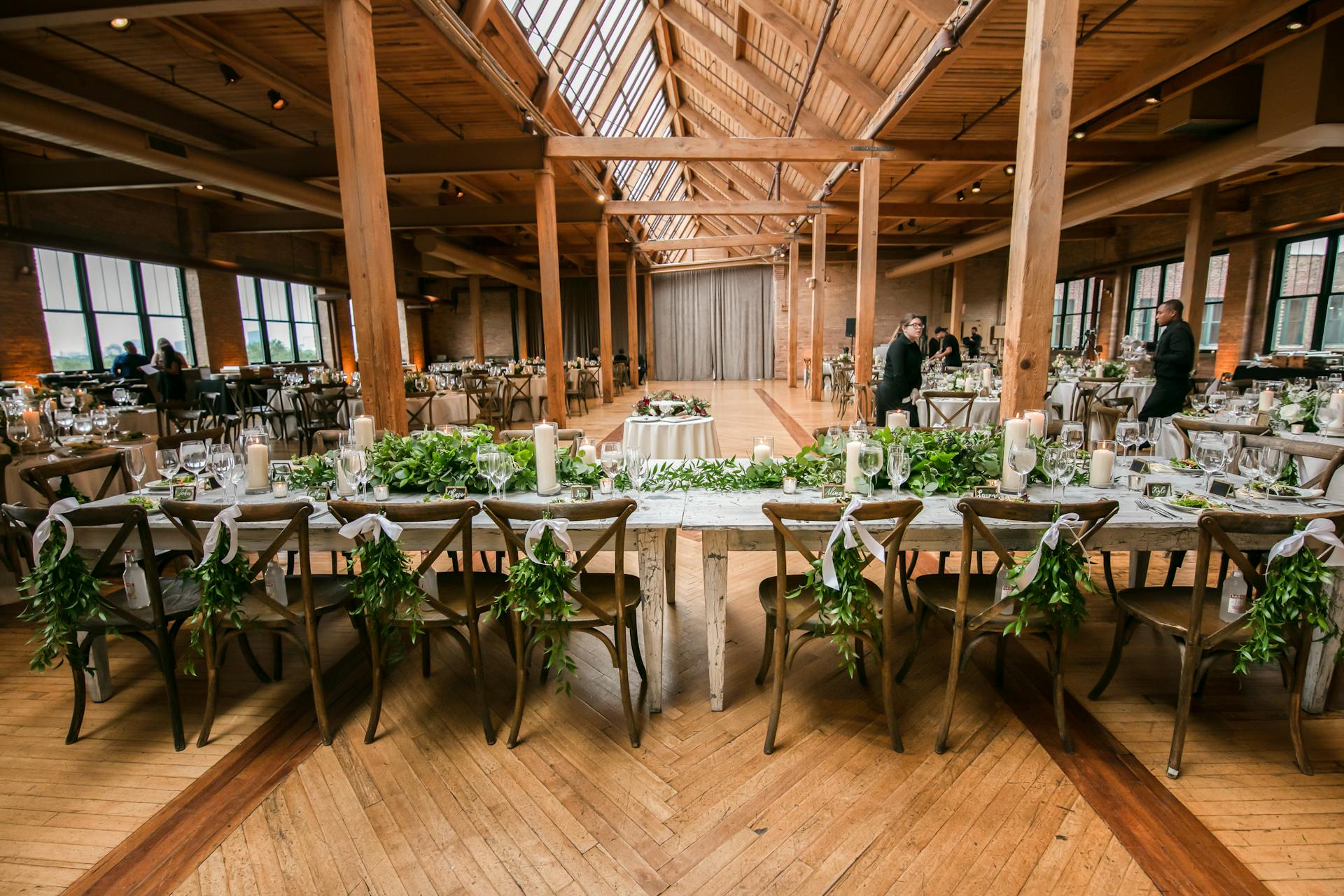 An elegant wedding reception venue with long wooden tables decorated with greenery, white candles, and neatly arranged glassware, set inside a spacious hall with tall wooden beams, skylights, and large windows.